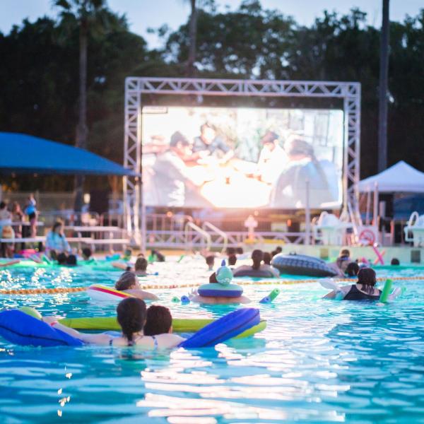 People floating on inflatable rafts in a swimming pool while watching a movie on a large outdoor screen at dusk, with palm trees in the background.