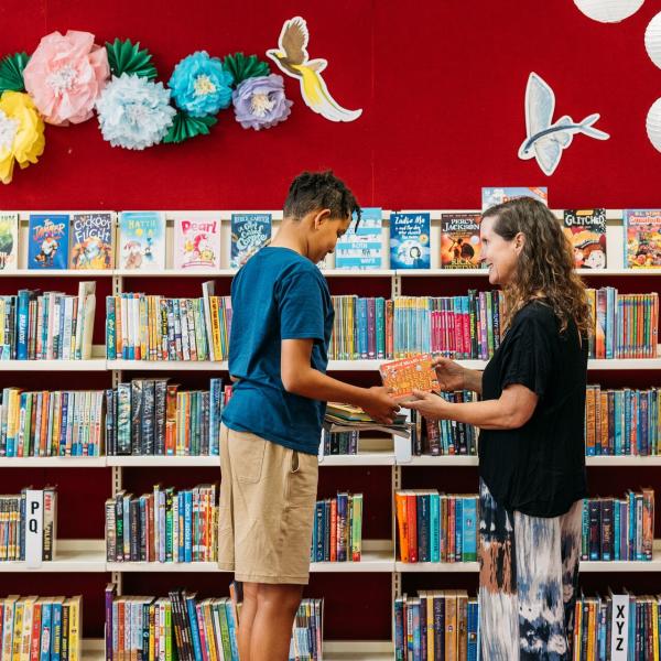 Women handing boy a book in front of a bookshelf 