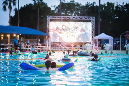 People floating on inflatable rafts in a swimming pool while watching a movie on a large outdoor screen at dusk, with palm trees in the background.