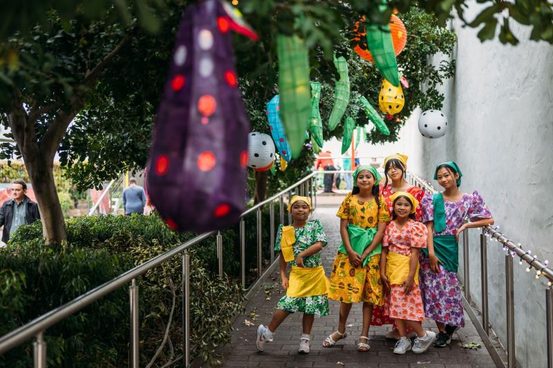 Group of children in colourful traditional outfits standing together on a garden path decorated with lanterns.