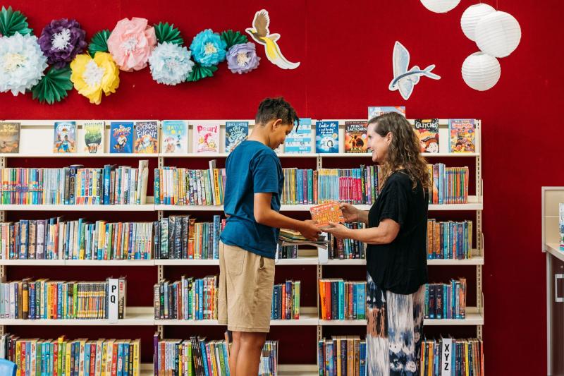 Women handing boy a book in front of a bookshelf 