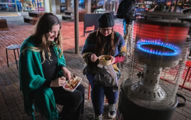 Two females sitting by a fire heater in The Levee with food rugged up