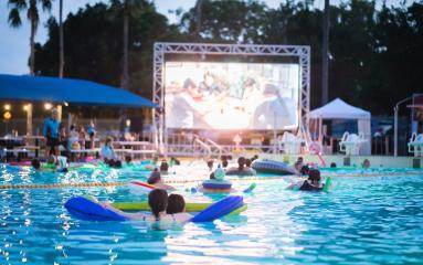 People floating on inflatable rafts in a swimming pool while watching a movie on a large outdoor screen at dusk, with palm trees in the background.