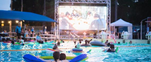 People floating on inflatable rafts in a swimming pool while watching a movie on a large outdoor screen at dusk, with palm trees in the background.